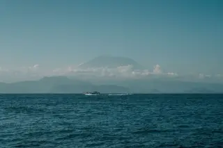 Illustration: A small boat cruises on the blue sea with a large mountain under clouds in the background.
