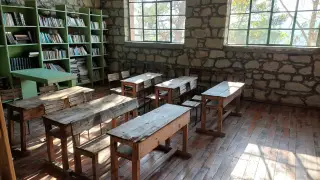 Illustration: Empty wooden desks and a bookshelf inside a rustic classroom with stone walls.