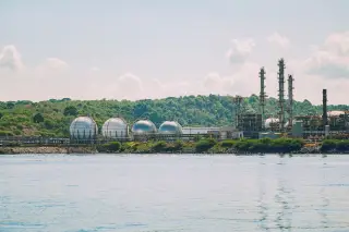 Illustration: Spherical industrial storage tanks and refinery towers situated on a green coastline by the sea.