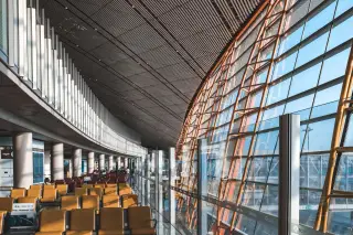 Illustration: Modern airport terminal interior with empty seating rows and large glass facade letting in sunlight.