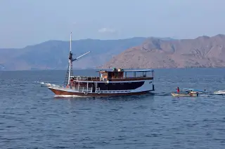 Illustration: A wooden phinisi boat sails across the sea with arid coastal hills in the background.