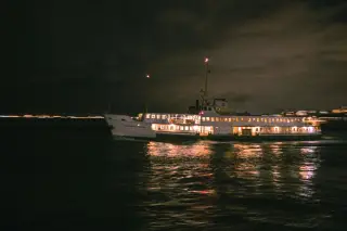 Illustration: An illuminated passenger ferry sailing at night across dark water.
