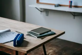 Illustration: Notebooks, papers, and a blue pencil case on a light wooden desk in an empty classroom.
