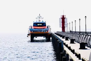 Illustration: A passenger ferry moored next to a long concrete pier with a red navigation tower.