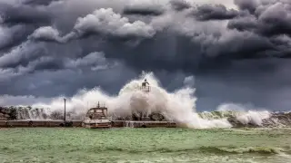 Illustration: Large ocean waves crash over a stone pier with a small boat and light beacon under dark storm clouds.