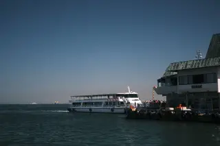 Illustration: A passenger ferry docks at a waterfront pier next to a terminal building under a clear blue sky.