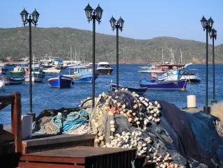 Illustration: Piles of fishing nets and buoys on a pier with boats moored in a calm harbor.