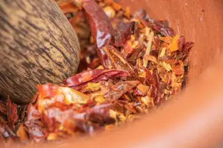 Illustration: Crushed dried red chili peppers and seeds being ground with a wooden pestle in a mortar.