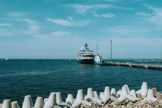 Illustration: A large vessel docked at a pier on open blue water behind concrete breakwater blocks.
