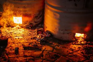 Illustration: Glowing embers and flames burn inside metal drum incinerator barrels on a stone ground.