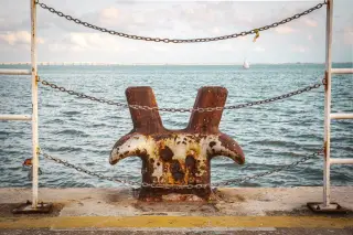 Illustration: A rusted mooring bollard on a dock framed by chains overlooking the sea.