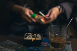 Illustration: A glass of iced coffee and an ashtray on a table with a person holding a cigarette pack in the background.