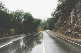 Illustration: A wet, paved mountain road winding through trees and misty hills under overcast skies.