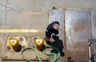 Illustration: Overhead view of a dock worker near yellow mooring bollards and green rope on a dock.