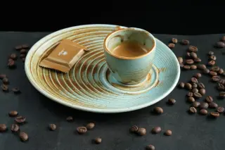 Illustration: An espresso cup on a patterned ceramic plate beside scattered roasted coffee beans on a dark surface.