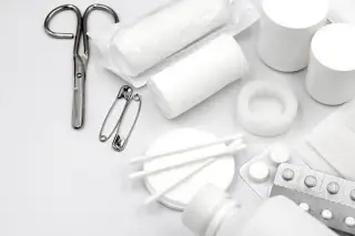 Illustration: A flat lay of white first aid supplies including bandages, scissors, pills, and cotton swabs.