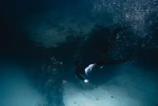 Illustration: A freediver in fins and swimwear swims underwater near a reef wall and sandy seabed.