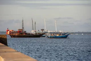 Illustration: Several wooden boats anchored on calm coastal water viewed from a stone pier.