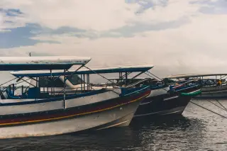 Illustration: Several motorized passenger boats moored side by side on the water under a cloudy sky.