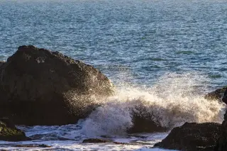 Illustration: Ocean waves crashing against dark coastal rocks with foaming spray in sunlight.