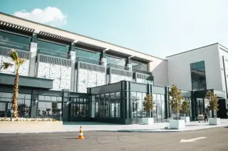 Illustration: Modern retail and commercial building with palm trees and a paved area under a clear sky.