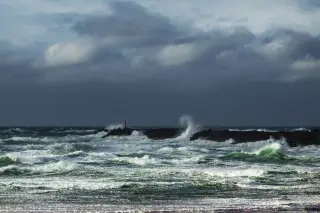 Illustration: Rough, stormy sea waves crashing against a dark breakwater under a cloudy sky.