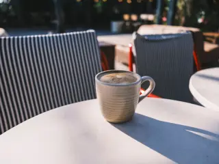 Illustration: A ceramic mug of coffee rests on an outdoor cafe table with striped chairs.