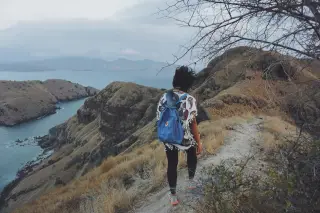 Illustration: A woman with a blue backpack walking along a grassy ridge overlooking coastal hills and the sea.