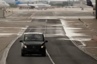Illustration: A black van drives on an airport tarmac with airplanes in the blurred background.